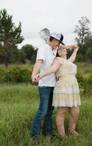 Playful couple dancing together in an outdoor field with pine trees in background.