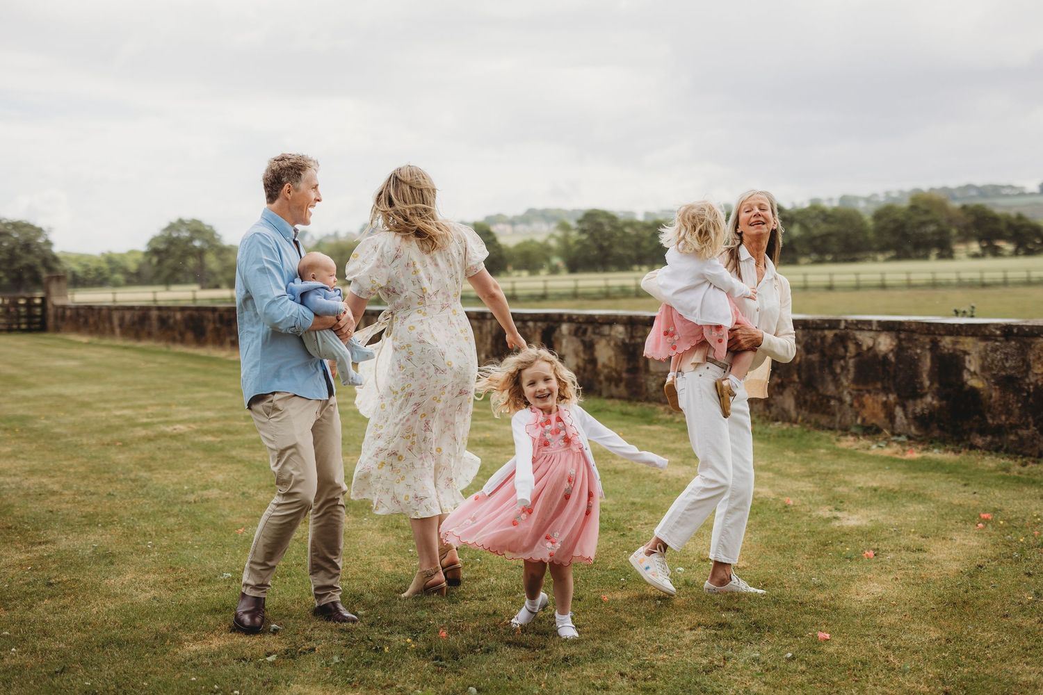 Group of people in casual spring outfits dancing joyfully together on a grassy field with wooden fencing in the background.
