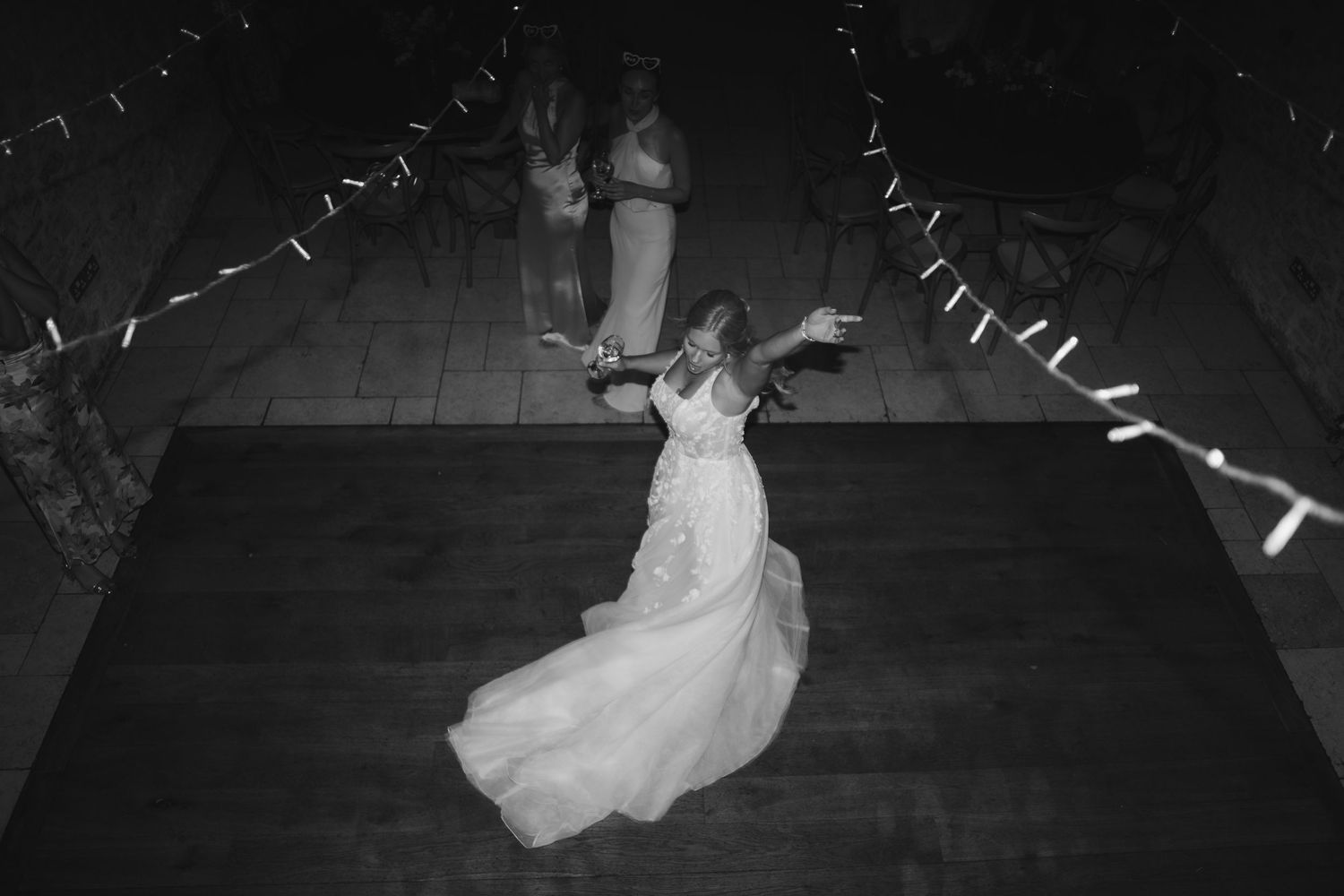 A black and white view of someone dancing in a flowing white wedding dress on a dark dance floor with festive decorations above.
