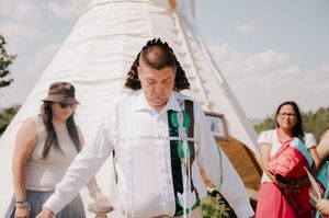 People in white clothing walk through a rustic outdoor ceremony setting with a white fabric canopy and wildflowers.