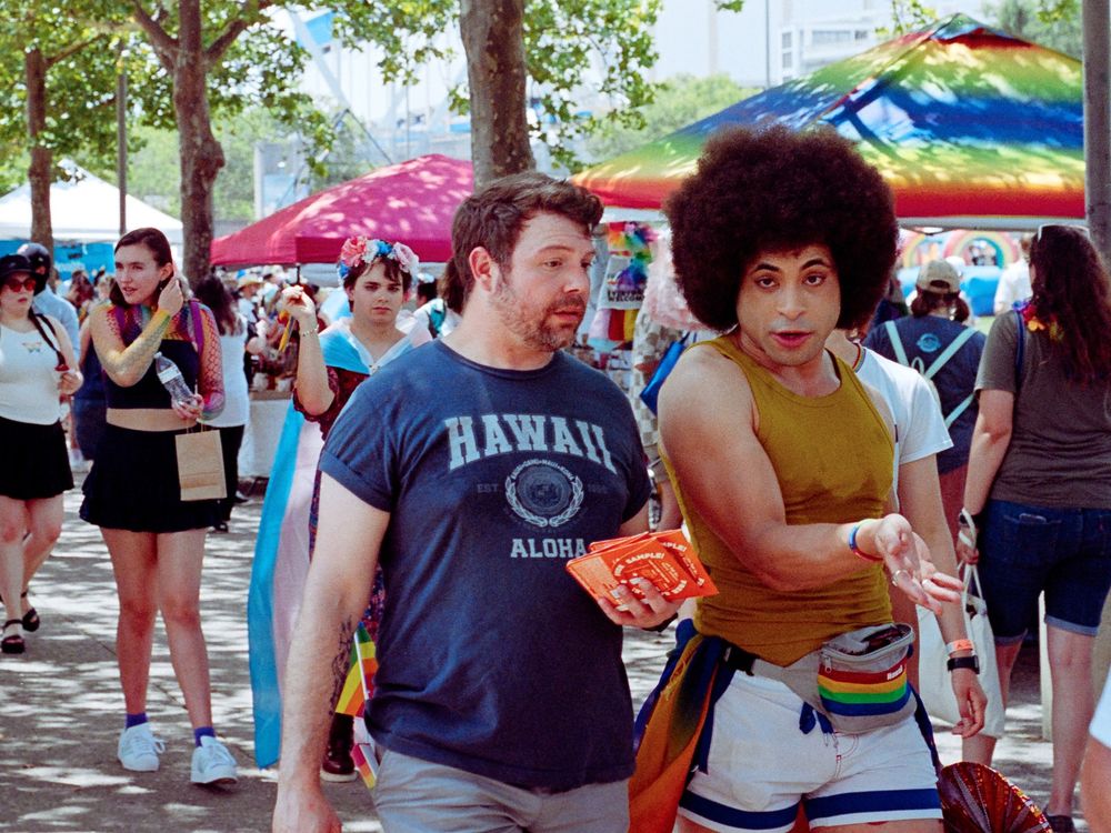 Festival goers enjoy a colorful outdoor event with umbrellas and rainbow decorations visible in the background.