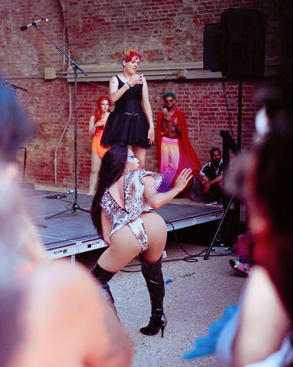 People dancing at an outdoor performance with brick wall backdrop and music equipment visible on stage.
