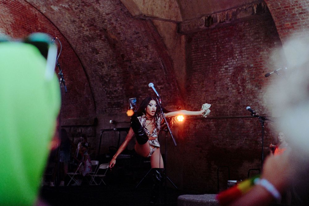 A performer in a theatrical costume holds up a glowing red orb while standing in a dark, brick-walled space.