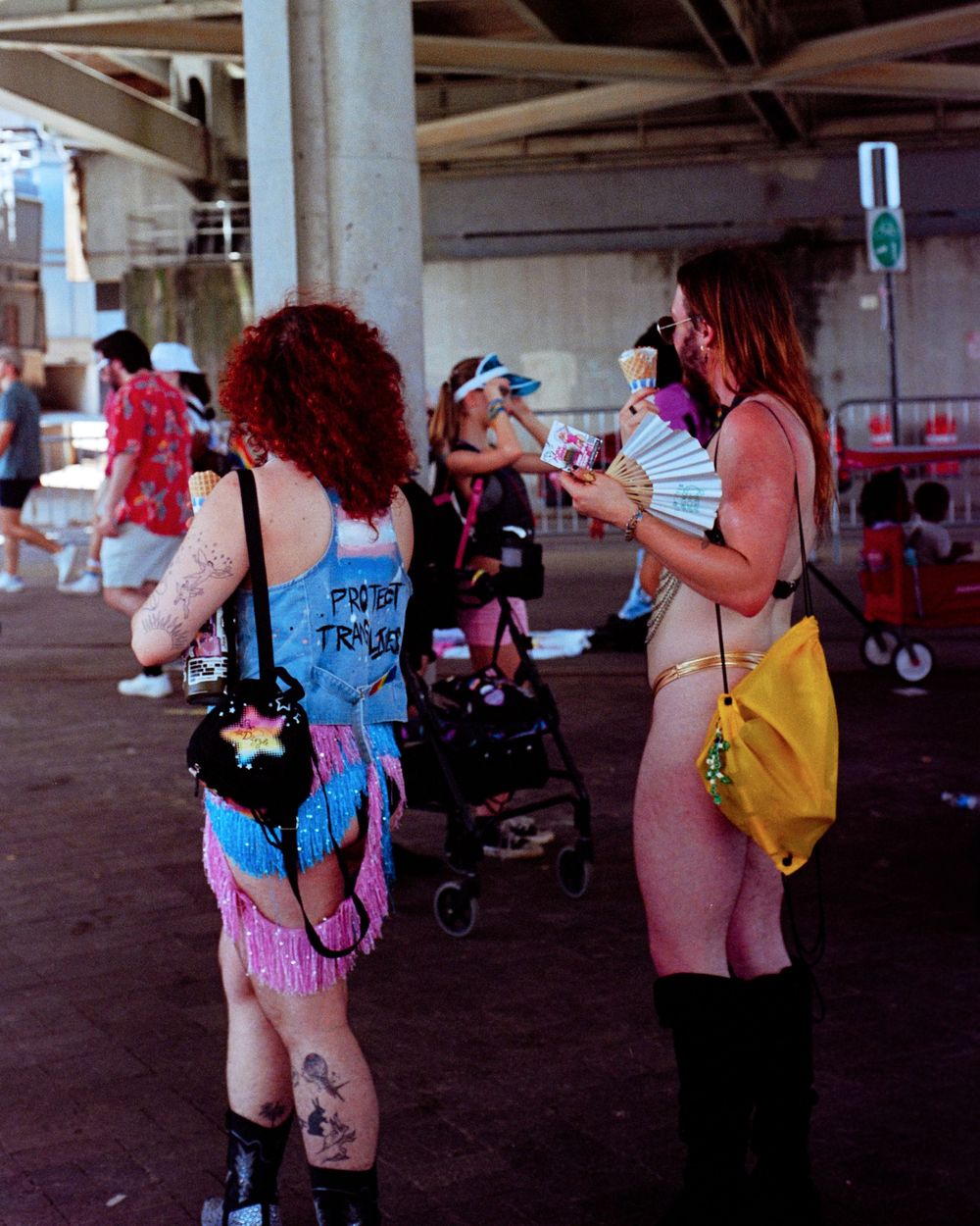 Two festival goers in colorful outfits chat under a concrete structure on a summer day.