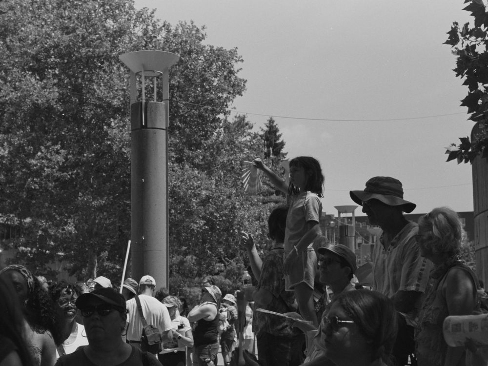 A black and white photograph shows a crowd gathered outdoors near trees and a cylindrical structure.