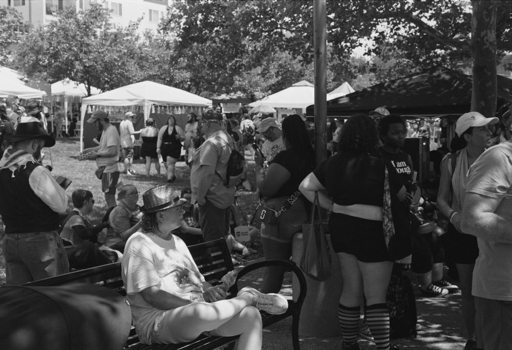Outdoor festival scene with people gathered under white tents and umbrellas on a sunny day.