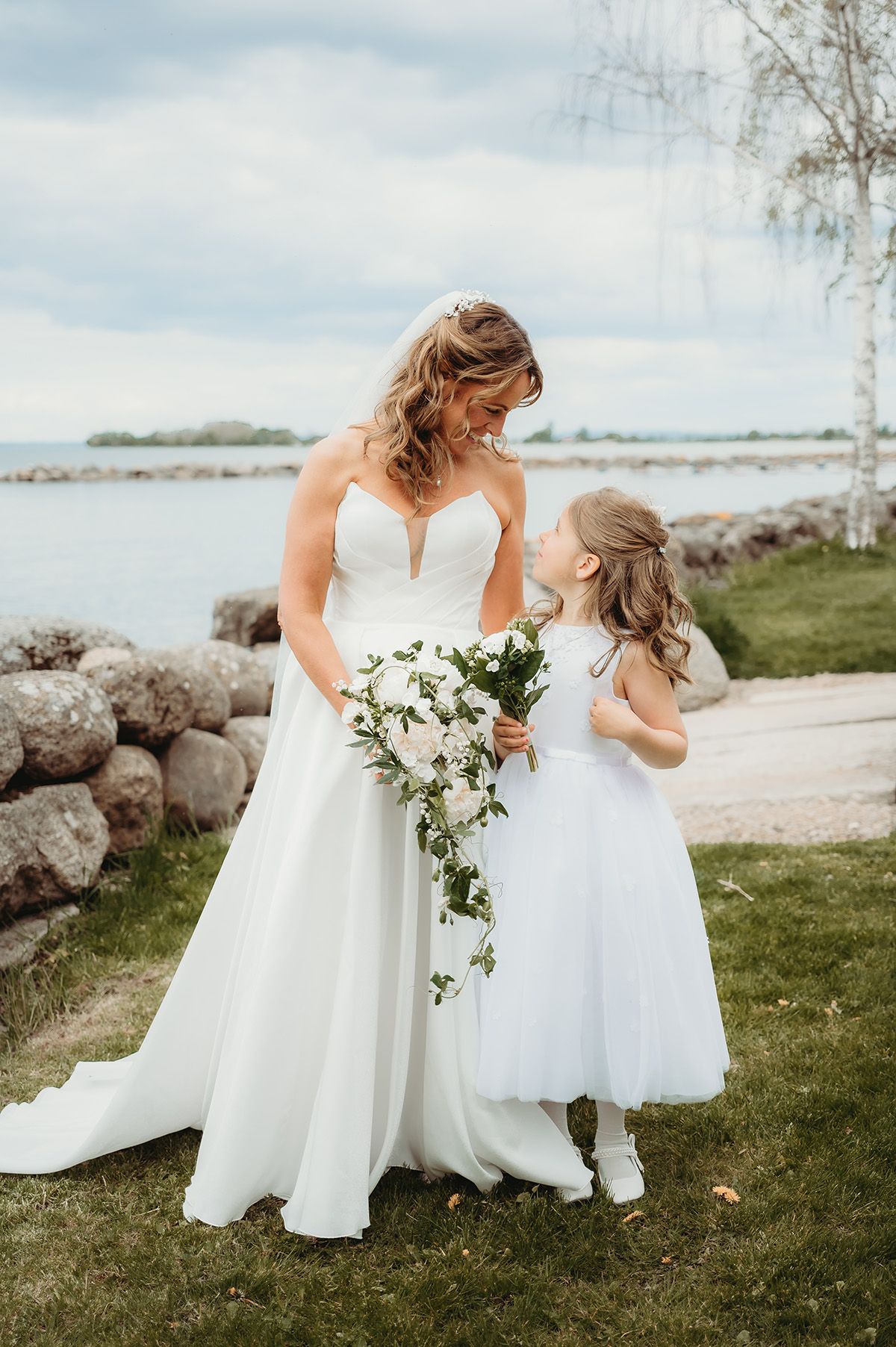Brud och brudnäbb ståendes vid Vätterns strand, de har vit klänningar och tittar på varandra. Bröllop i Vadstena, fotograf Josefin Landhagen