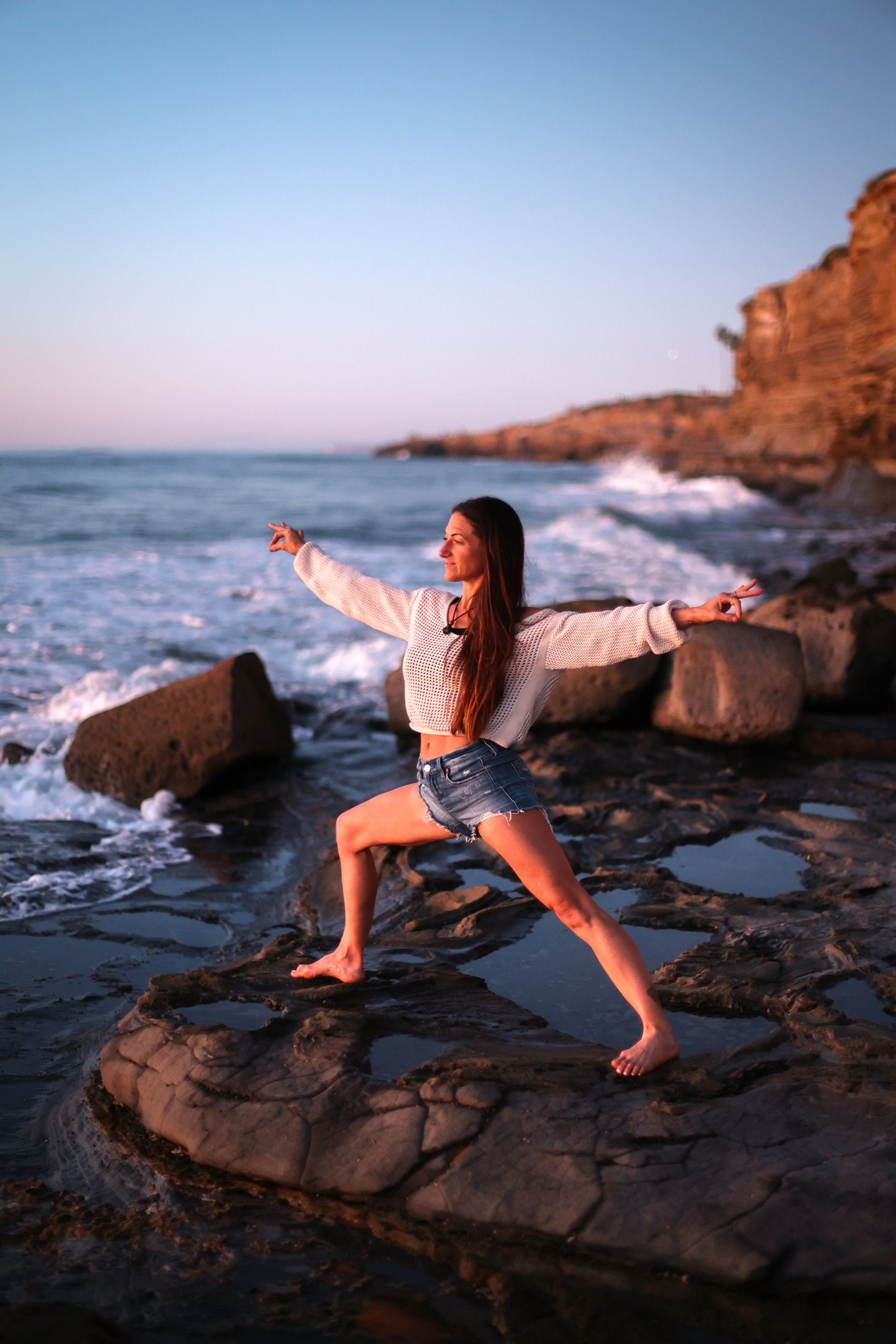 Woman striking yoga pose on coastal rocks with ocean waves at dusk.