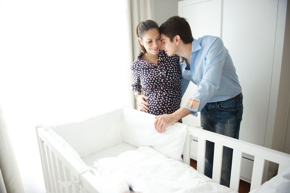 Expecting parents set up a white wooden crib while preparing a nursery in a bright room. Captured by GTA maternity photographer Phoebe Lee.