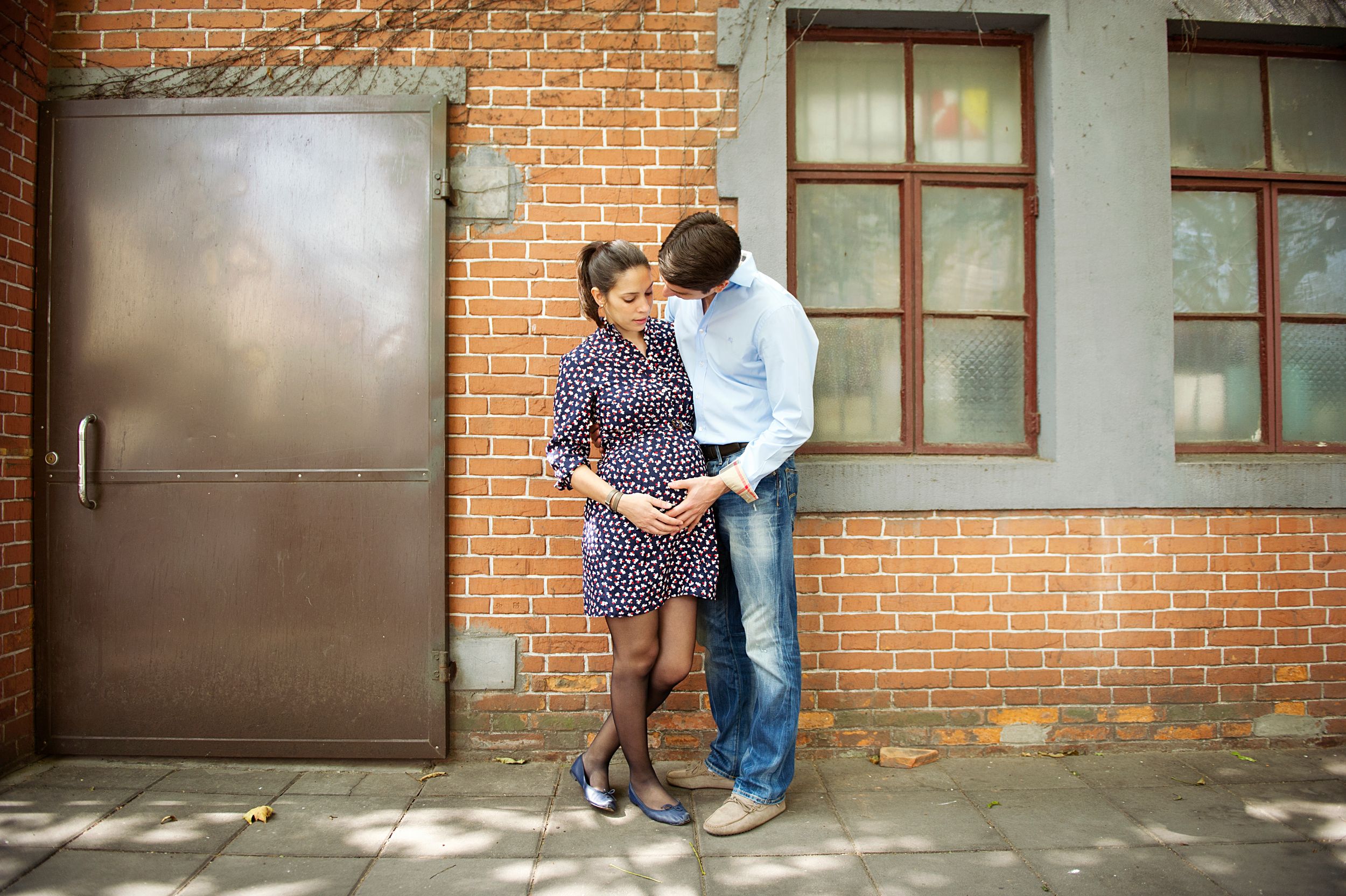 A couple shares an intimate moment against a brick wall with a metal door and windows in the background. Toronto Maternity Photography