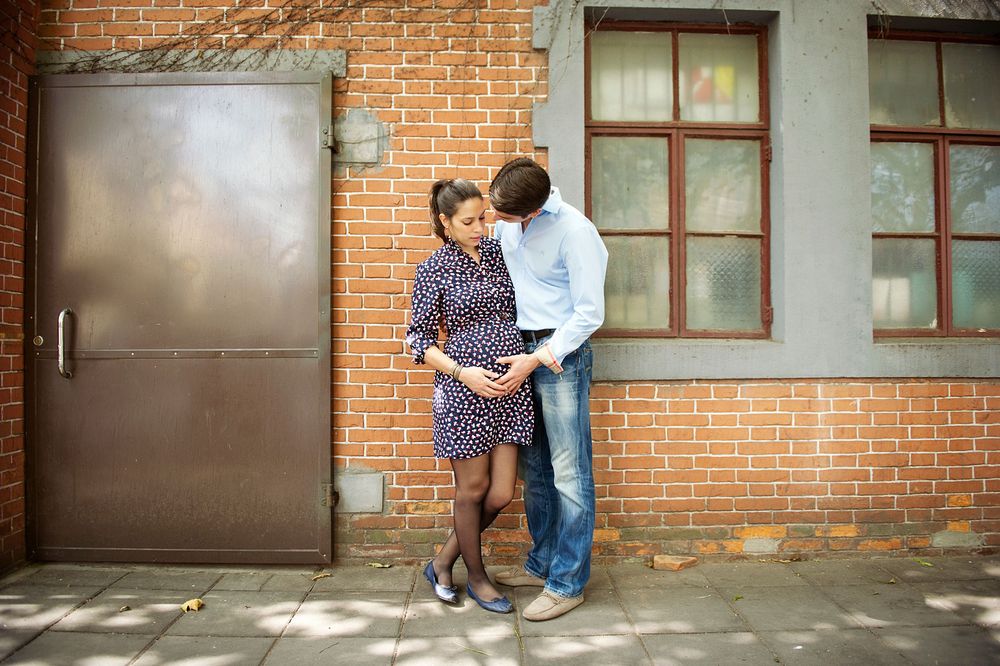A couple shares an intimate moment against a brick wall with a metal door and windows in the background. Toronto Maternity Photography