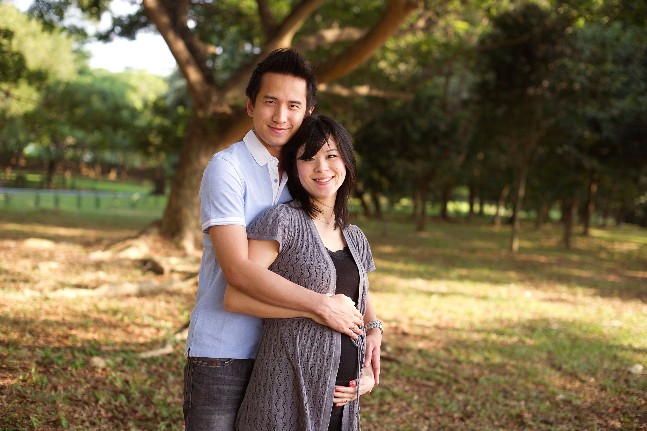 An expecting couple embraces in a warm outdoor setting with sunlit trees and natural scenery in the background. Captured by Phoebe Lee Photo