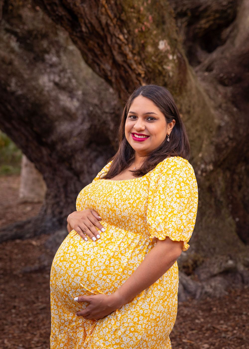 Expectant mother in yellow floral dress poses against rustic tree backdrop.