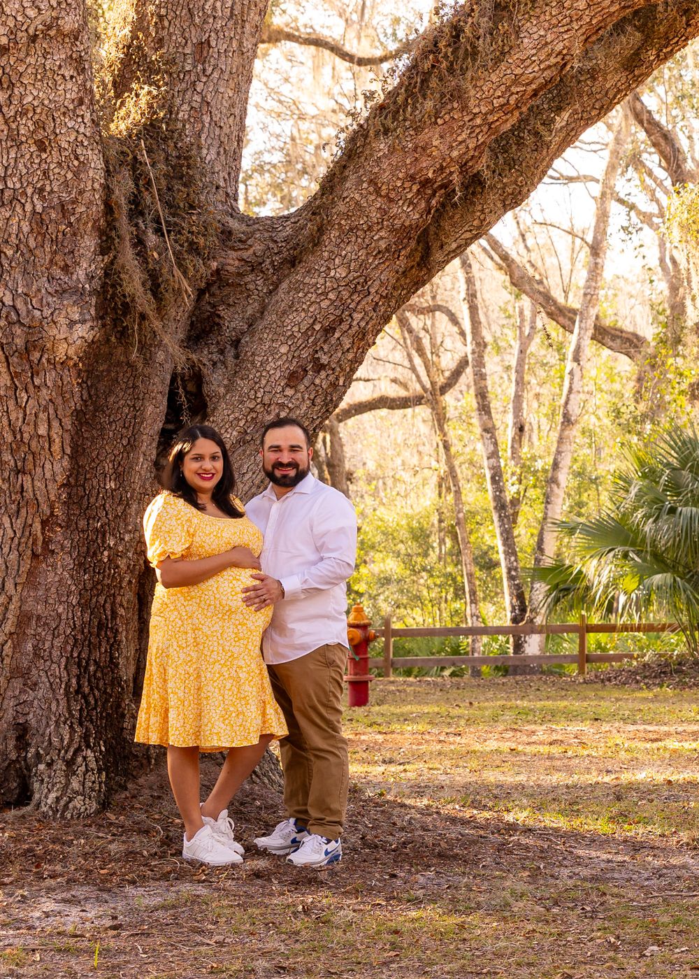 Couple poses together beneath massive oak tree in park setting.