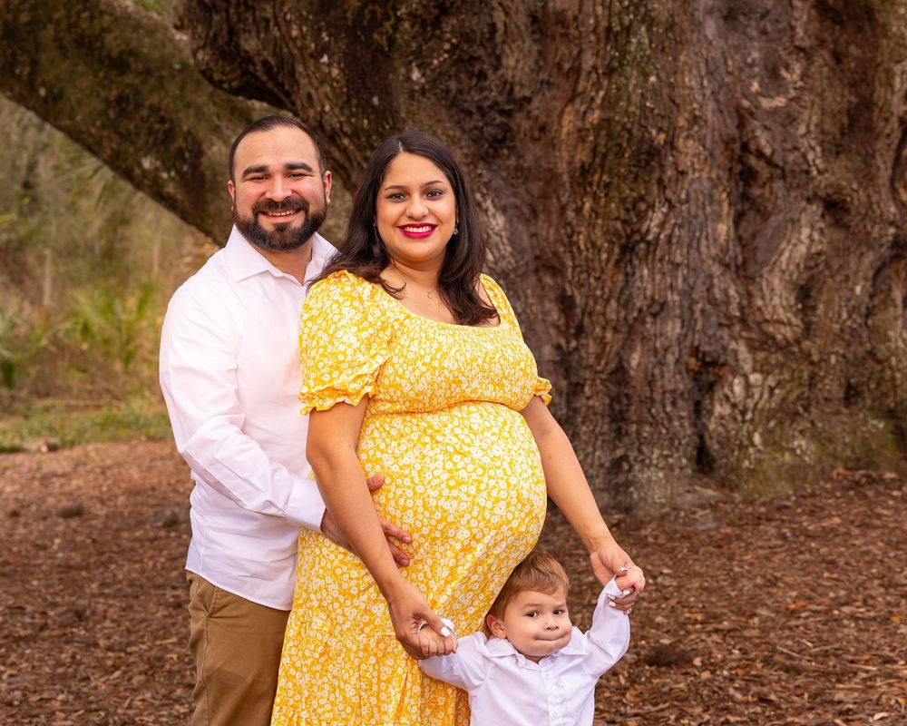 Family poses together in front of large tree trunk with person in yellow floral dress and white shirt.