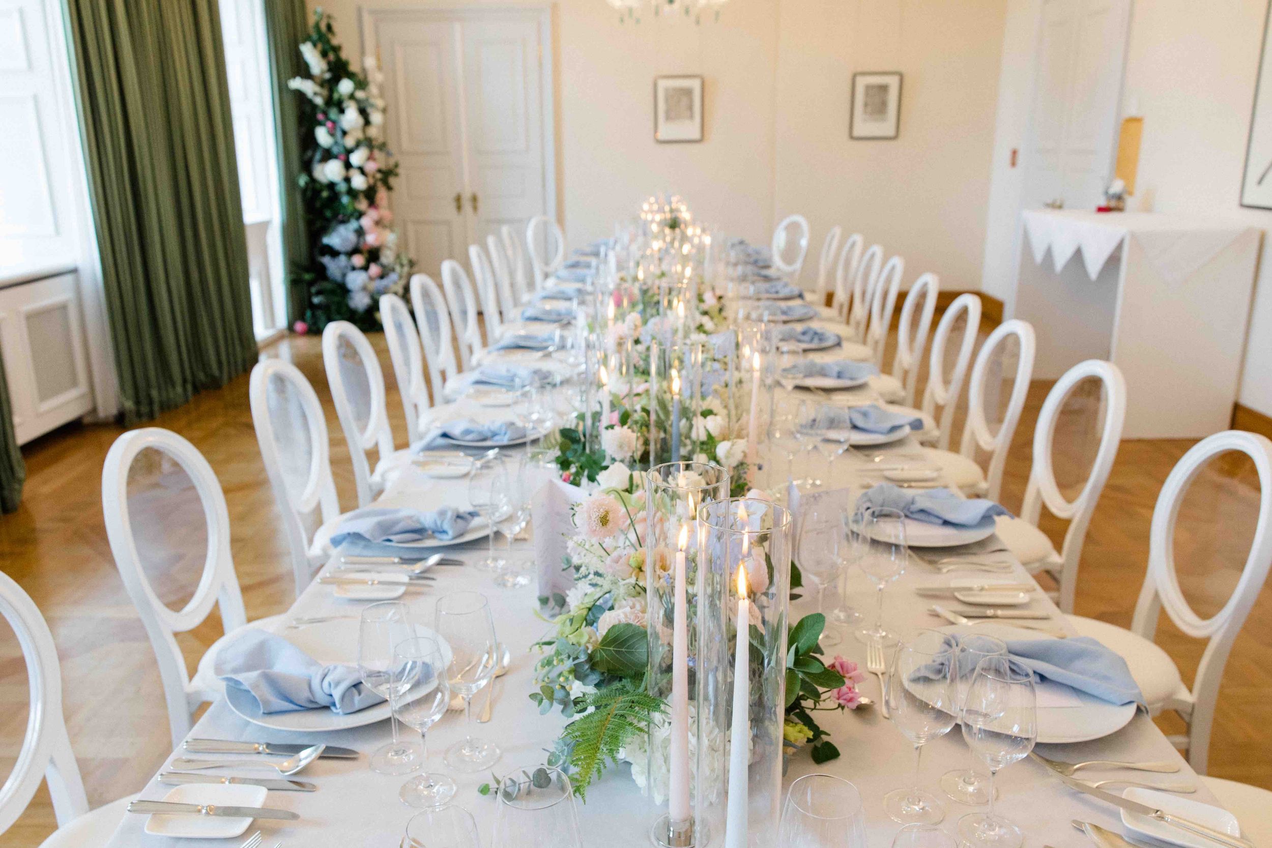 Elegant long dining table with white chairs and crystal candelabras set for formal wedding reception in bright room.