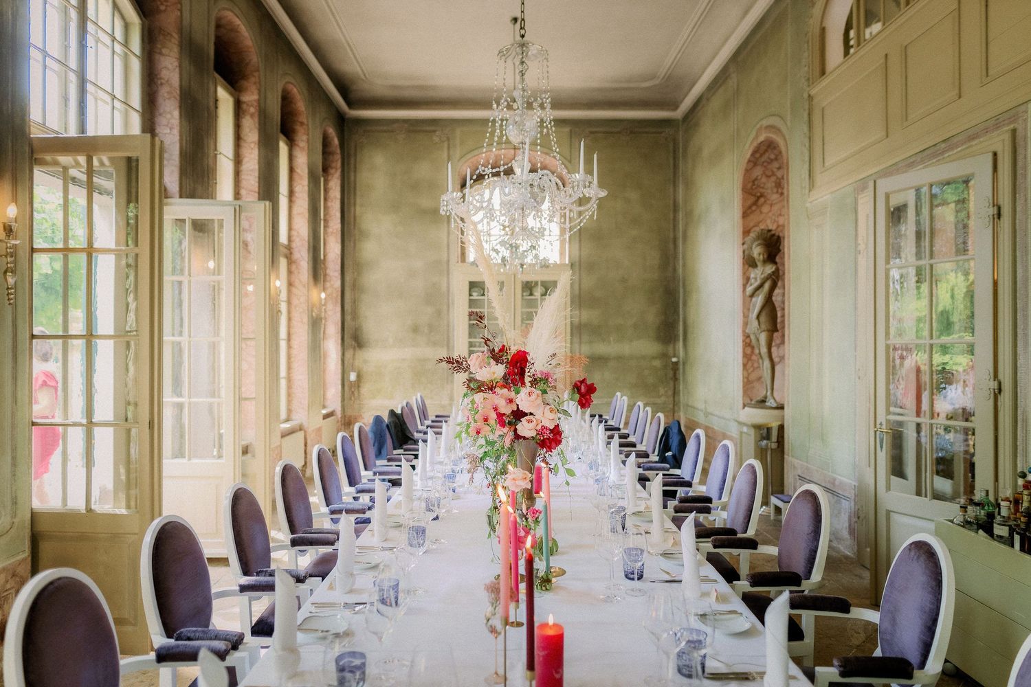 Elegant dining room with sage green walls, crystal chandeliers and pink floral centerpieces set for a formal event.