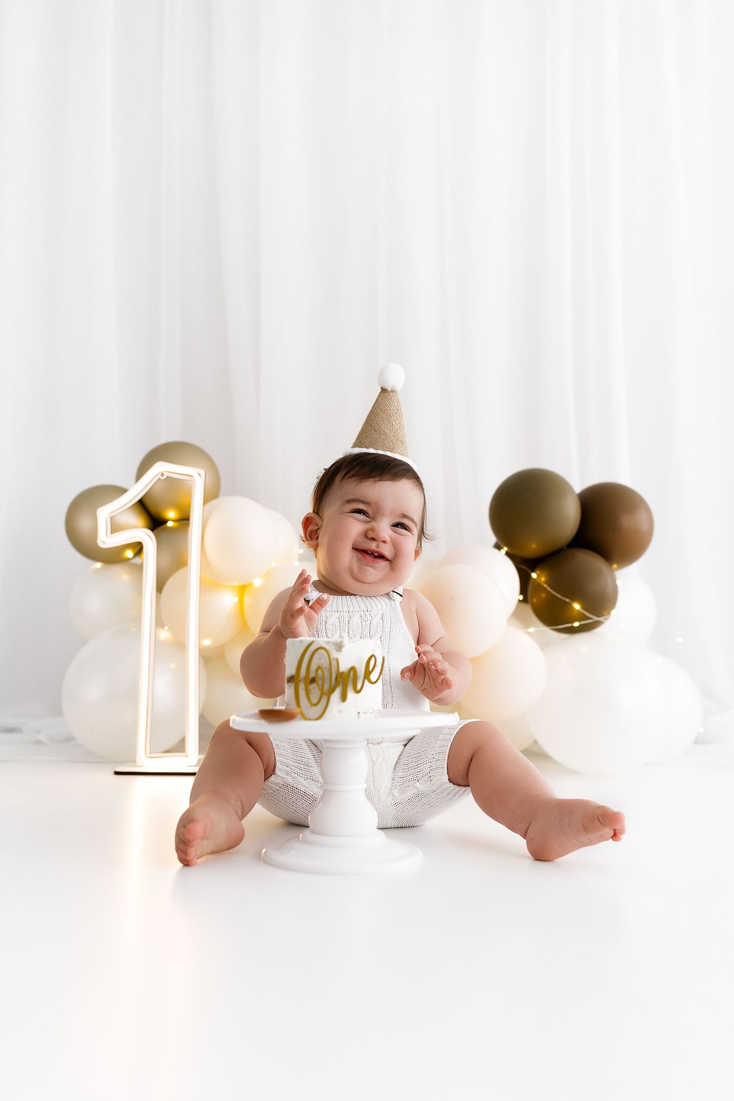 A smiling baby wearing a party hat and a white outfit sits on the floor with balloons and party decor.