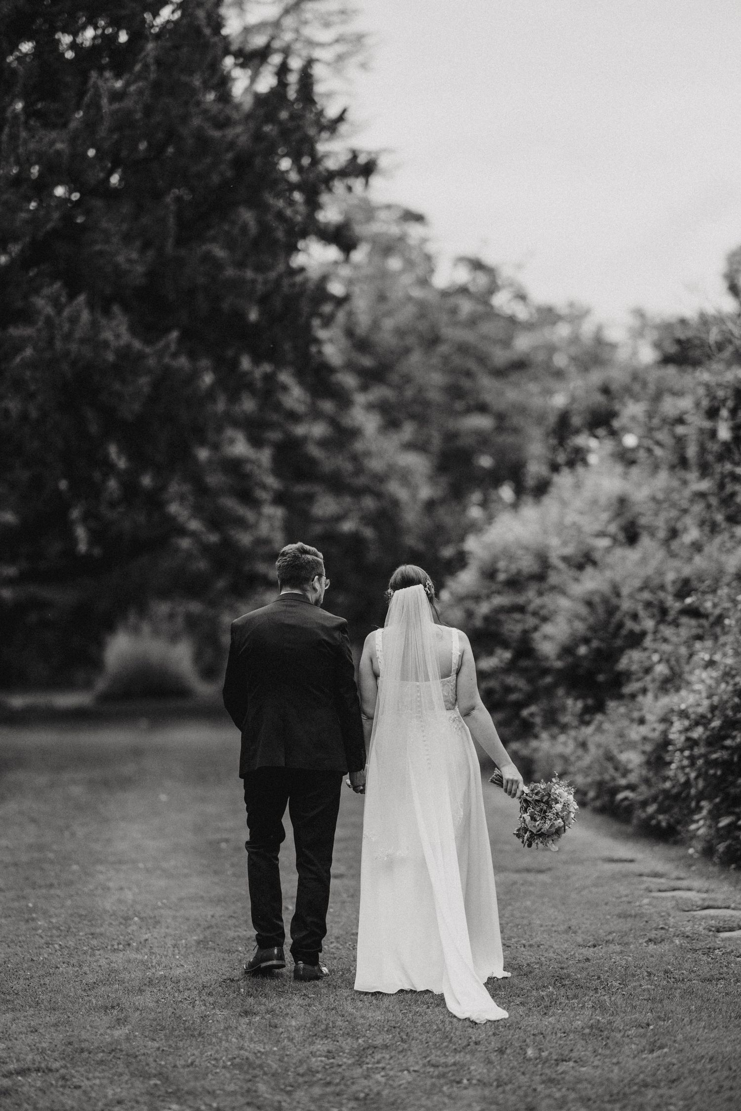 Black and white photo of a bride and groom walking together on a path through a garden in romantic candid style.