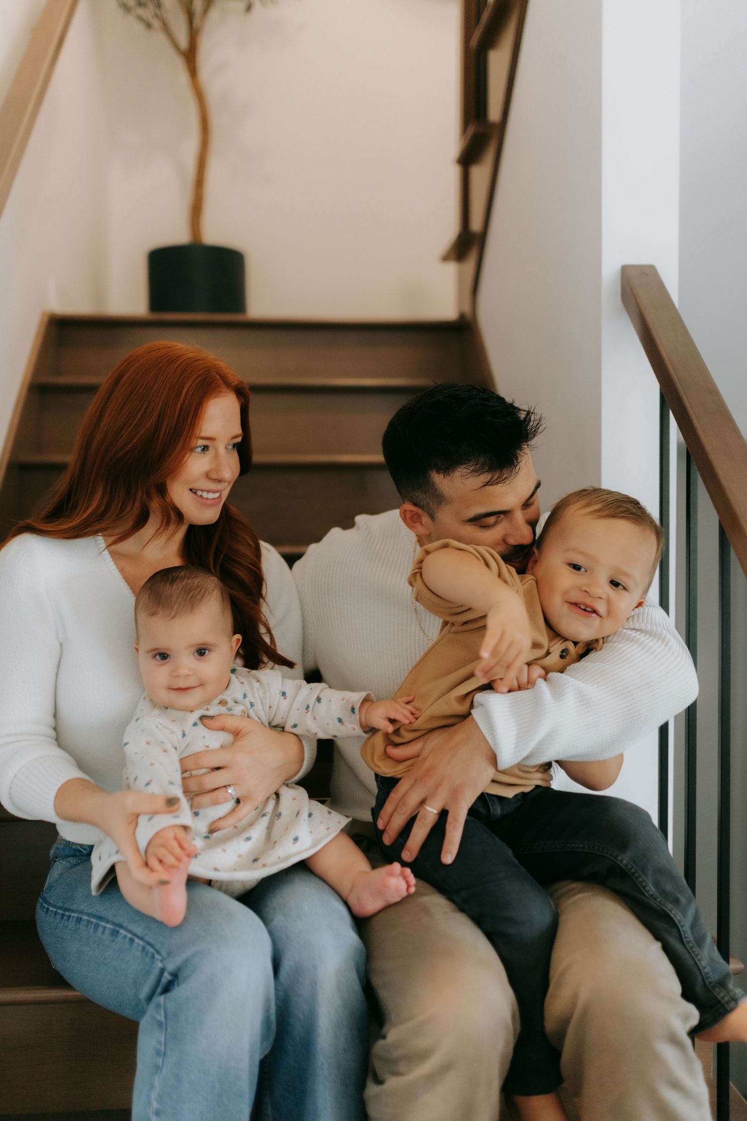A happy young family with two babies sits on wooden stairs, mom with red hair holding infant, dad kissing toddler.