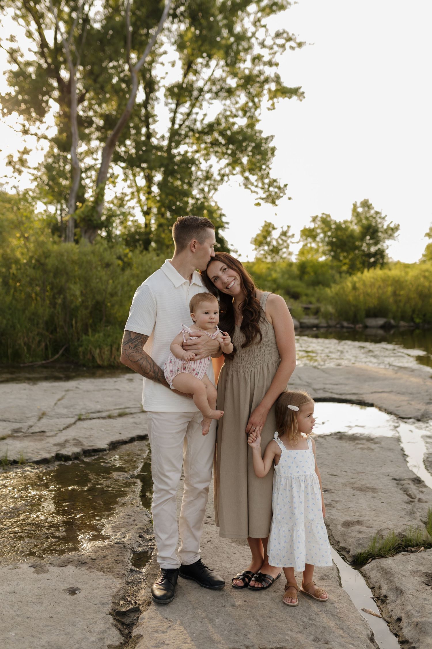 A happy young family with two babies sits on wooden stairs, mom with red hair holding infant, dad kissing toddler.