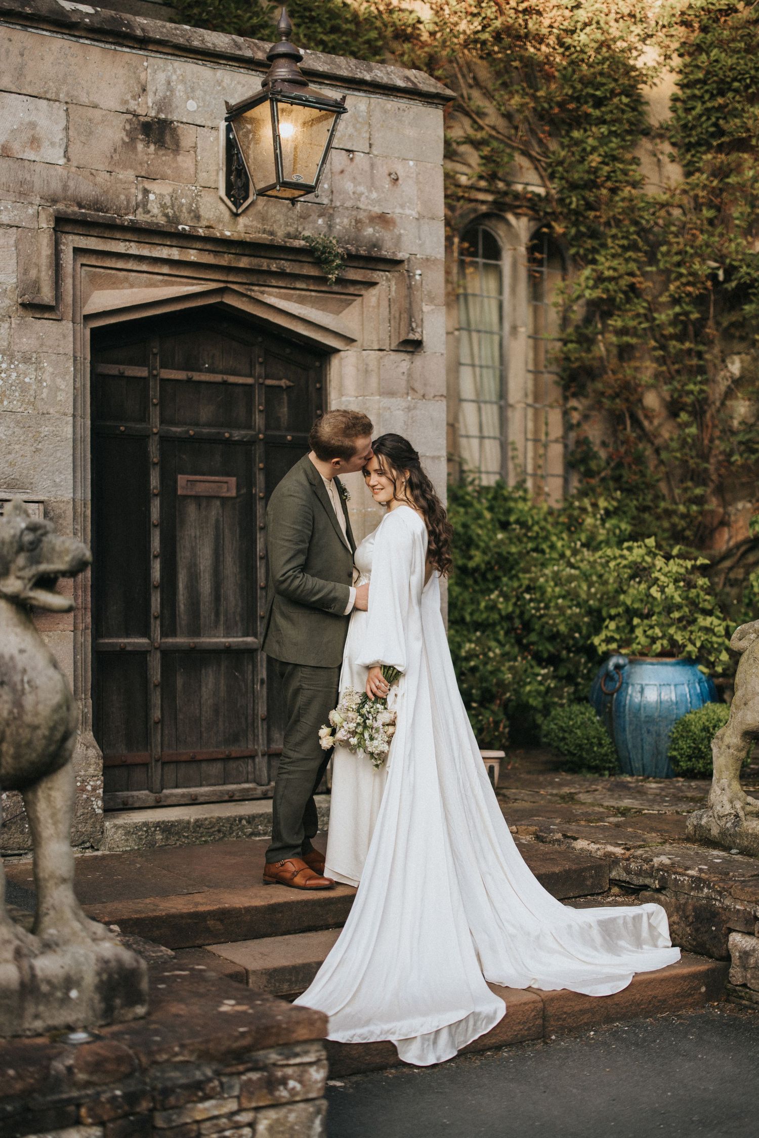 A couple shares an intimate moment in vintage wedding attire outside an old stone building with a rustic wooden door.