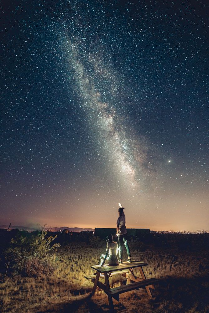 A glowing Milky Way galaxy over joshua tree stretches across the desert landscape while headlamps create light trails in the sand.