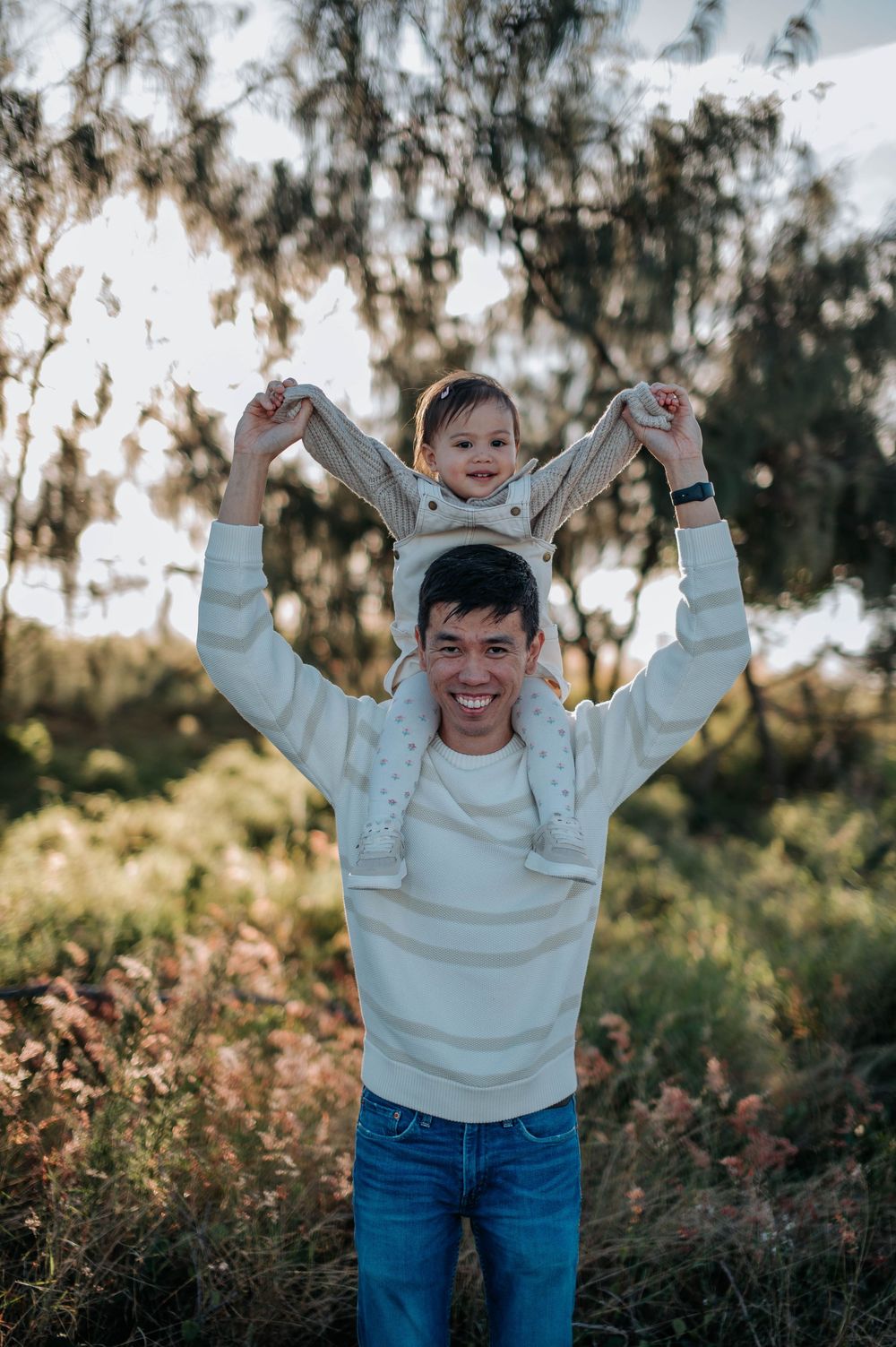 Joyful parent wearing white sweater lifts small child high in their arms outdoors against sunlit trees at golden hour.