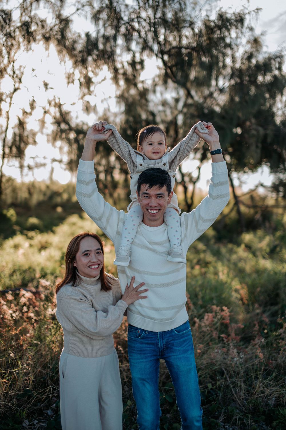 Two children in matching cream sweaters stand together in an outdoor setting with trees at sunset, sharing joyful expressions.