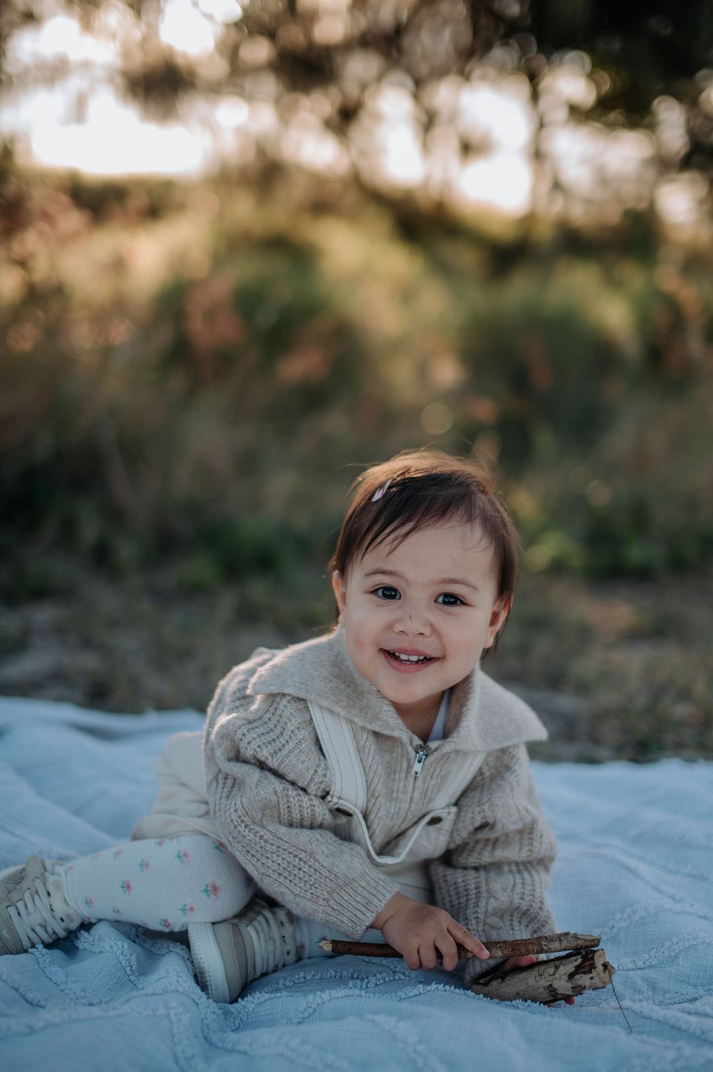 A small child wearing a cozy gray knit sweater lies on a white blanket in an outdoor setting amid soft bokeh lighting.