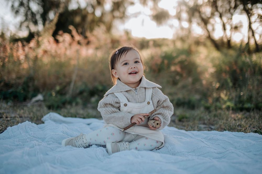 A small child sits on a white blanket in an outdoor setting during golden hour sunlight.
