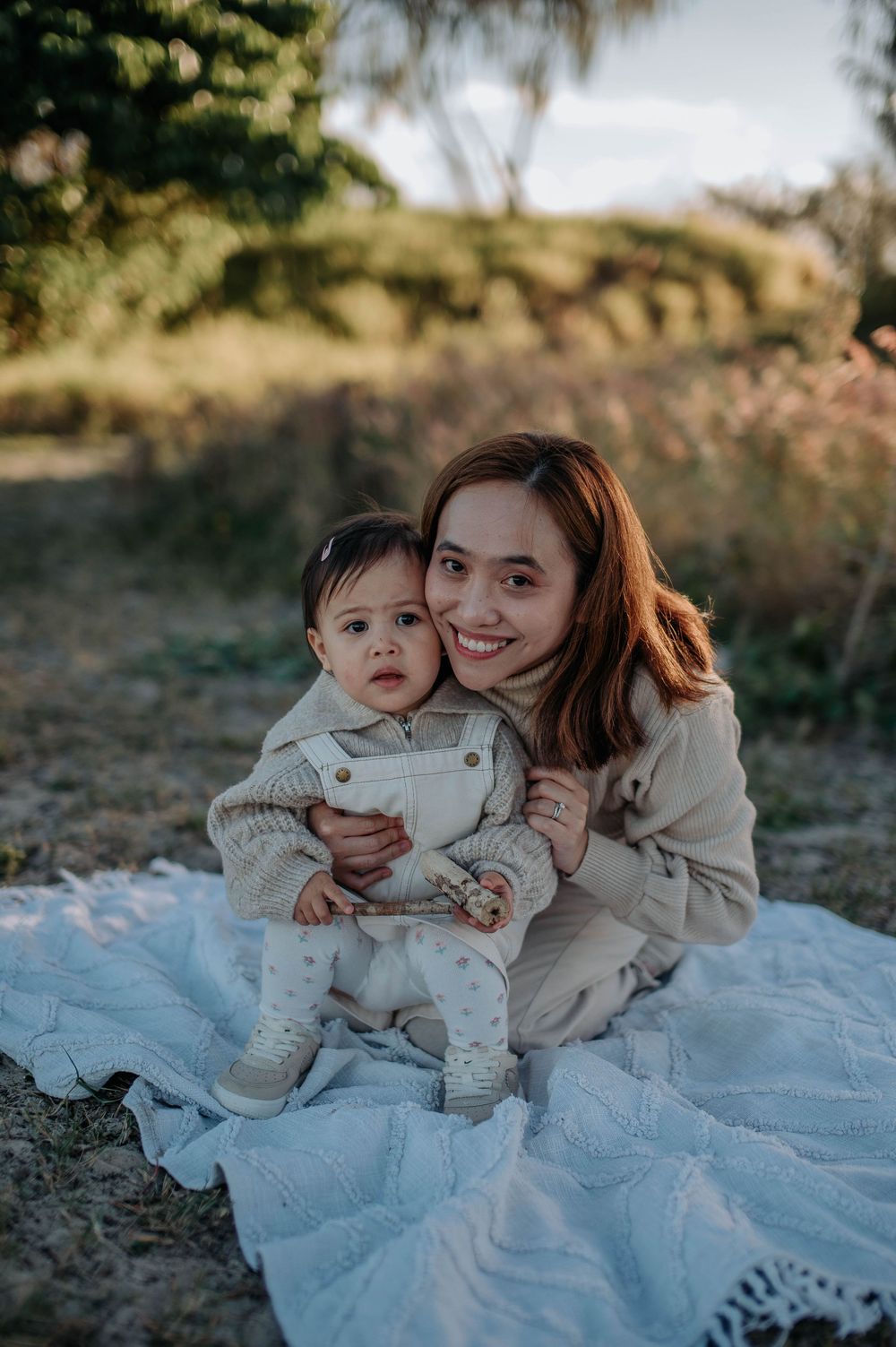 A woman and a baby sit on a light blue blanket outdoors at sunset, sharing tender moments together.