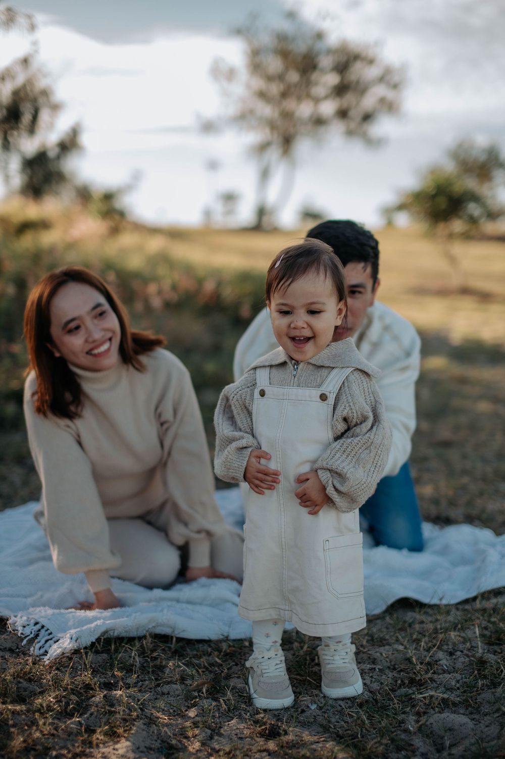 A toddler laughs joyfully while standing on a picnic blanket outdoors with their family on a sunny afternoon.