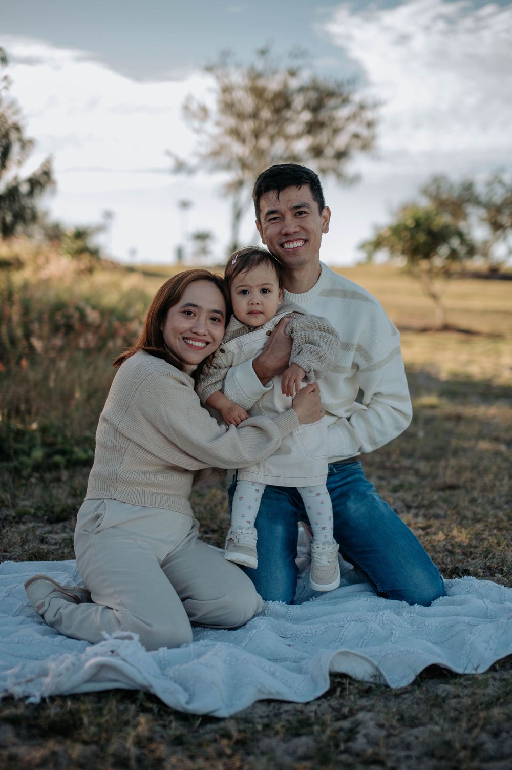 Young family in coordinating cream sweaters having a cozy outdoor photoshoot on a blanket in nature.