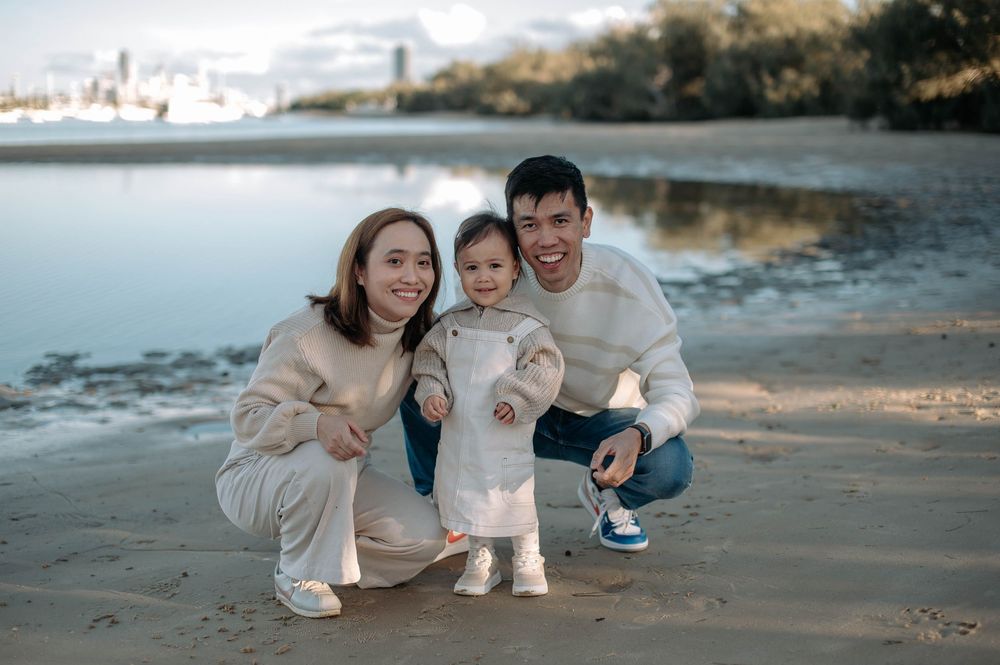 A small family crouches together on a beach at sunset with mountains and water in the background.