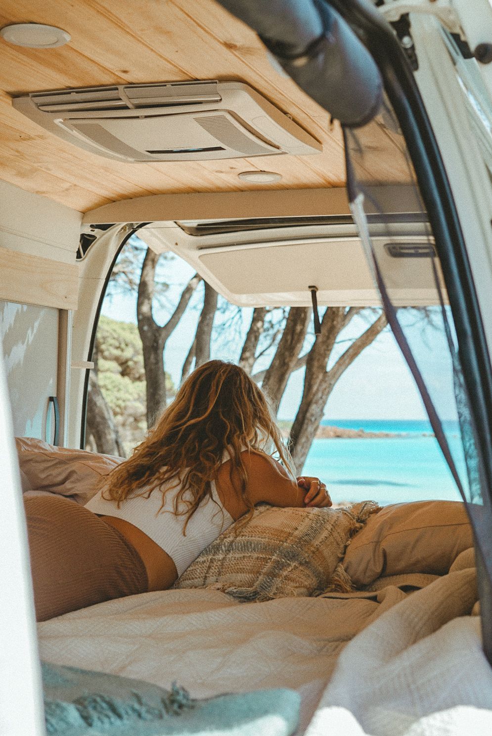 View through camper van window of ocean scenery with cozy bed setup.