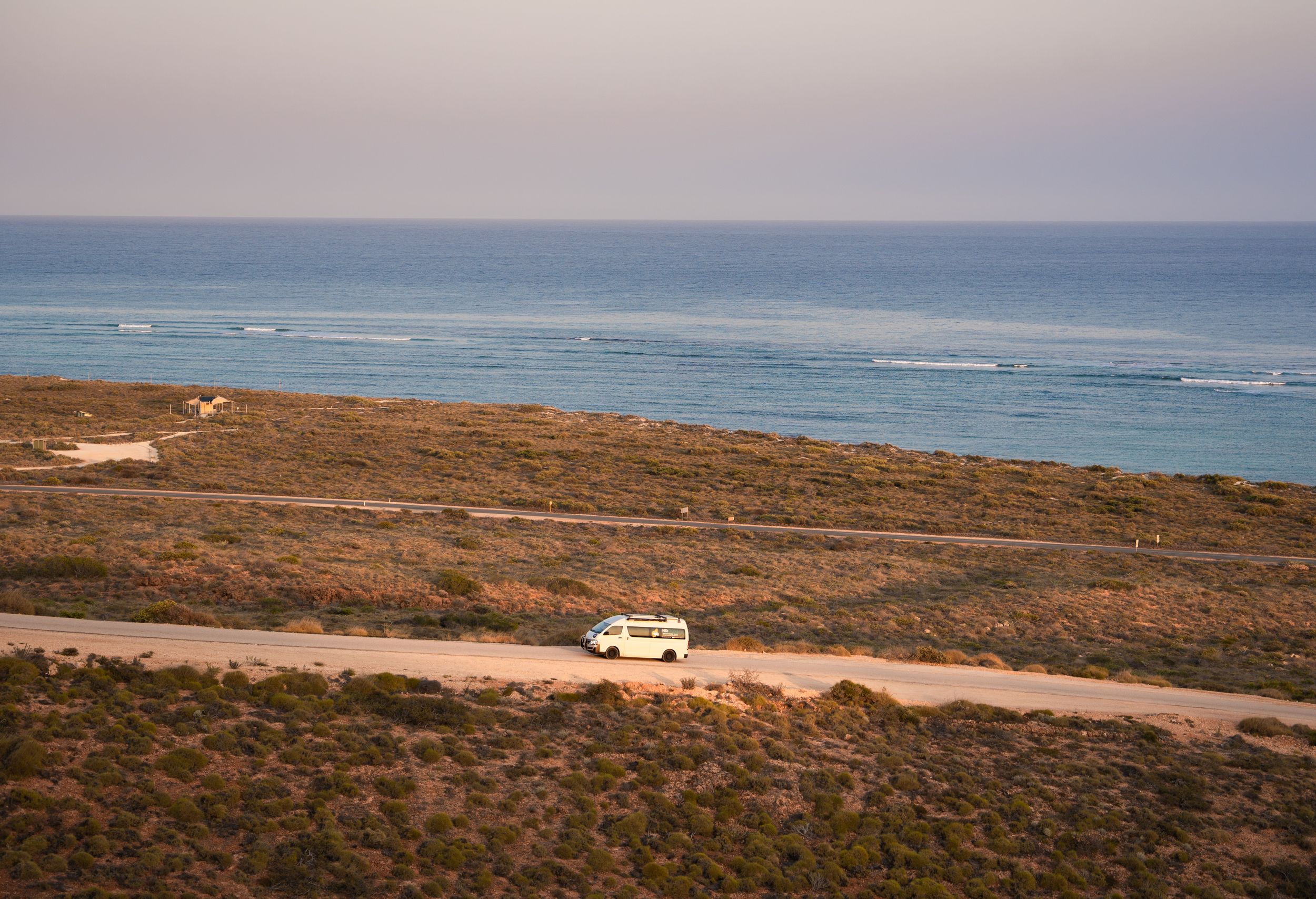 Aerial view of a white van driving along a coastal dirt road with ocean views.