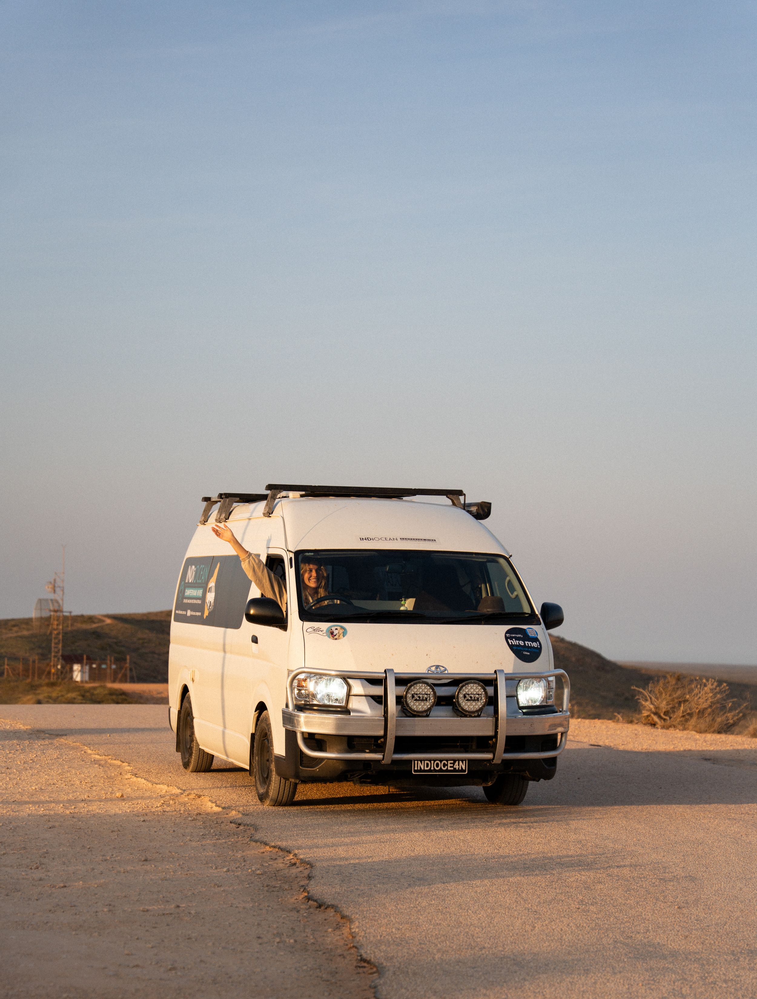 A white camper van with roof rack parked on a coastal dirt road at dusk.