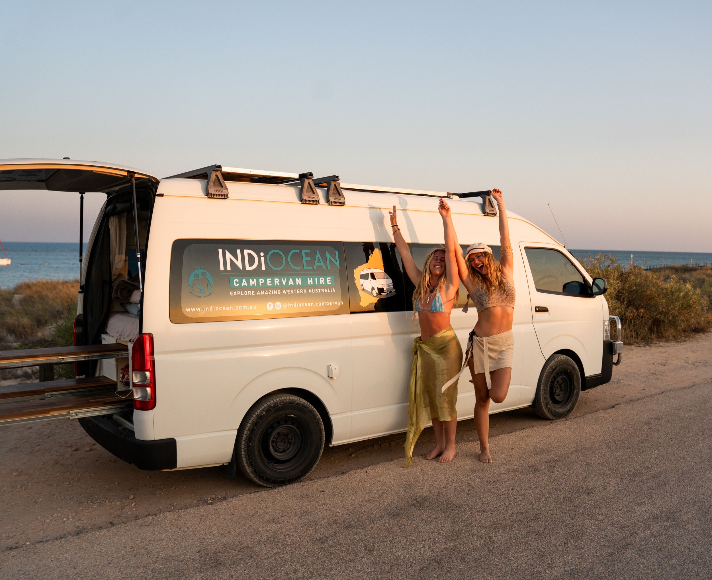 Two people standing by a white campervan with 'INDI' branding against an ocean backdrop.