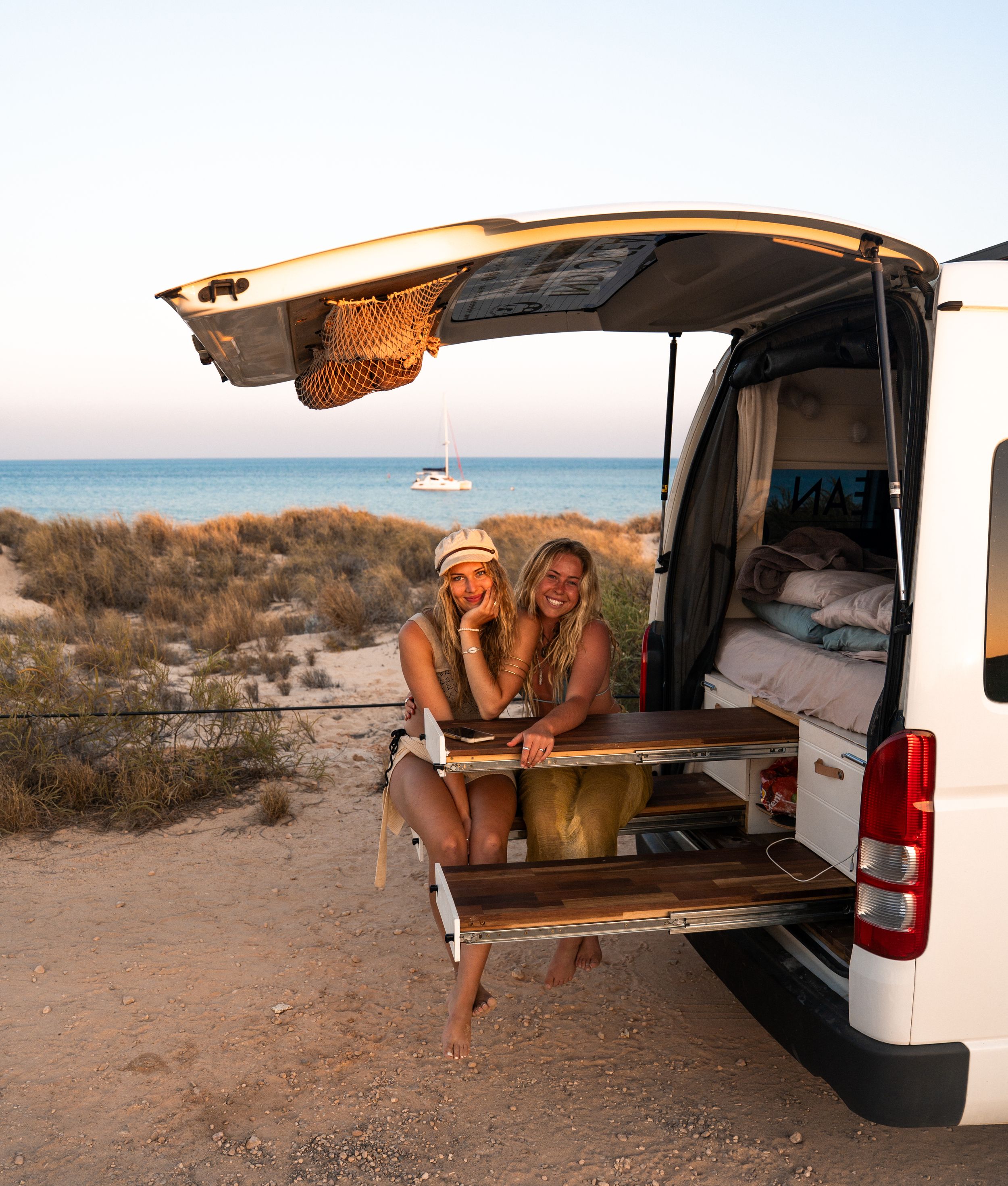 A camper van parked on a coastal cliff with people relaxing in the open back door at sunset.