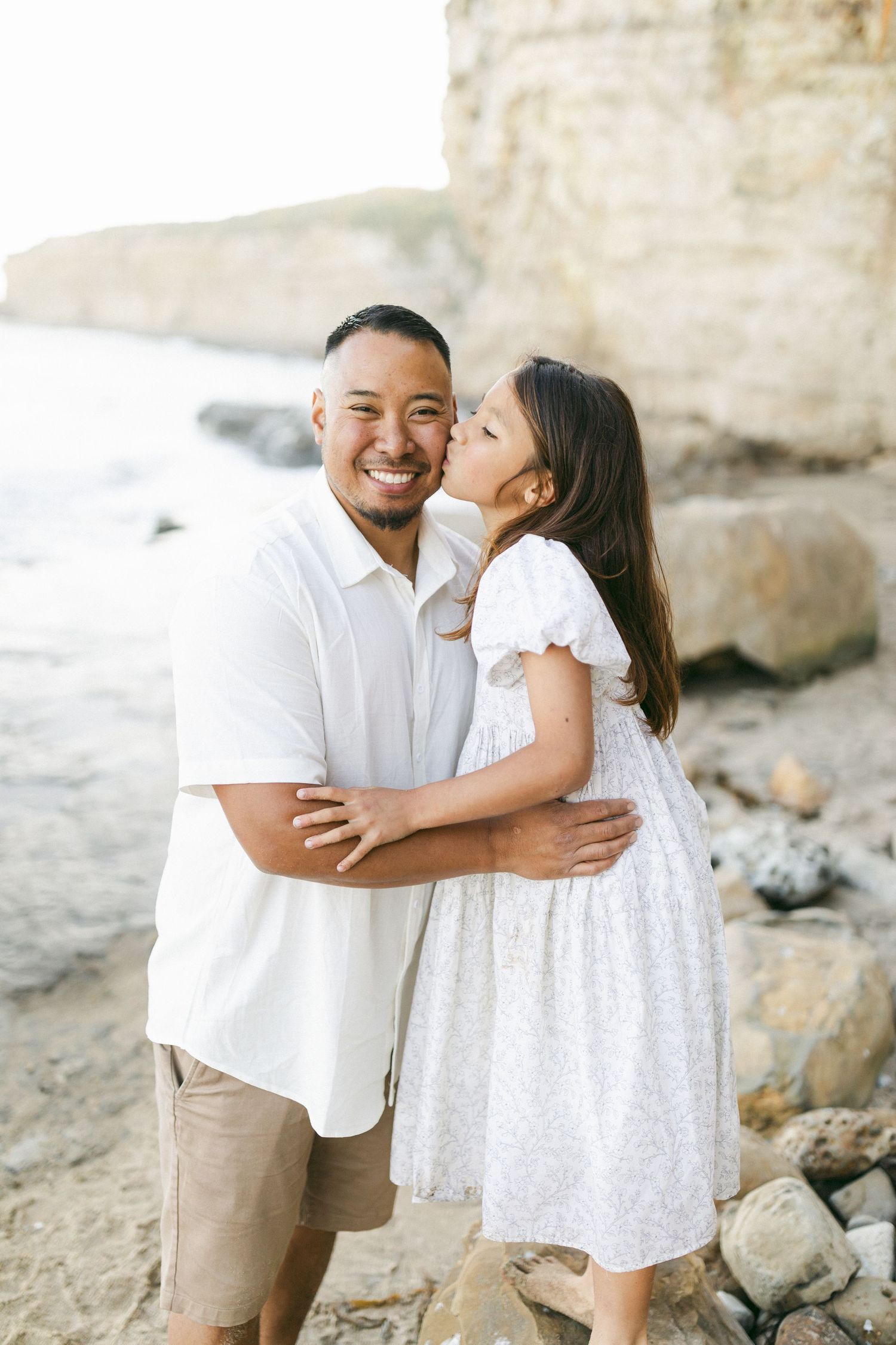 A couple in white outfits embracing on a rocky beach during a romantic photoshoot against coastal cliffs film by Chelsee Rawe .