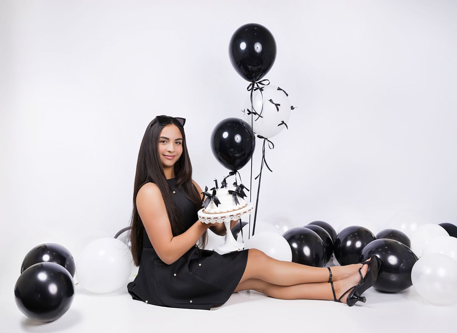 Woman in black dress sits on white floor surrounded by elegant black balloons in studio setting.