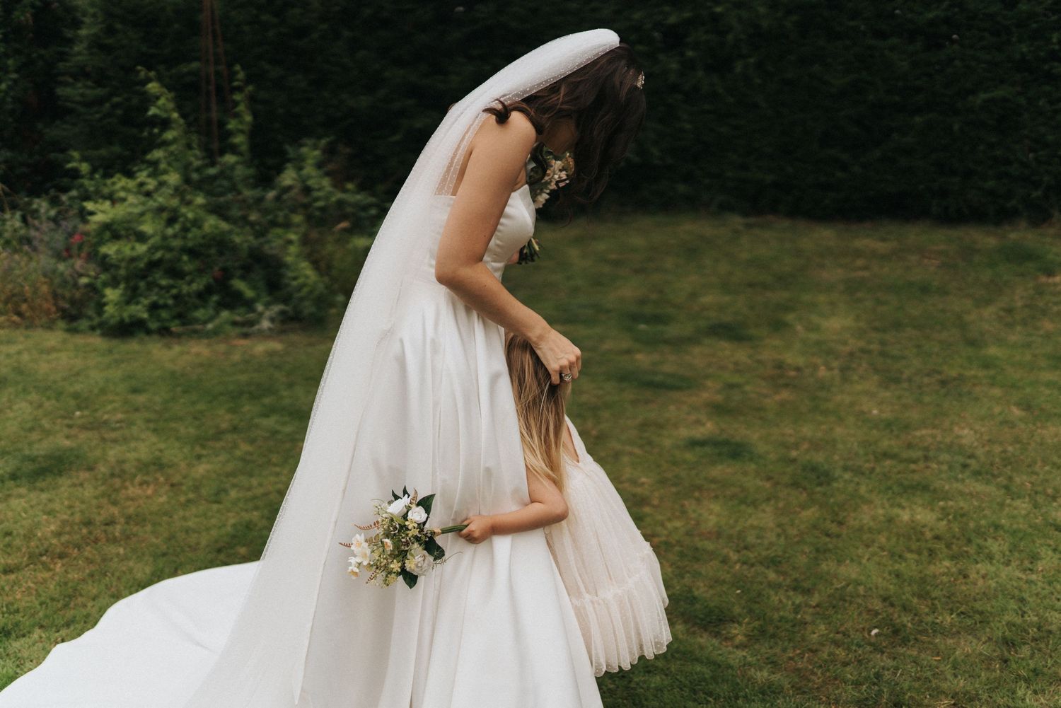 A tender moment between figures in white wedding attire holding a bouquet of flowers on a green lawn.