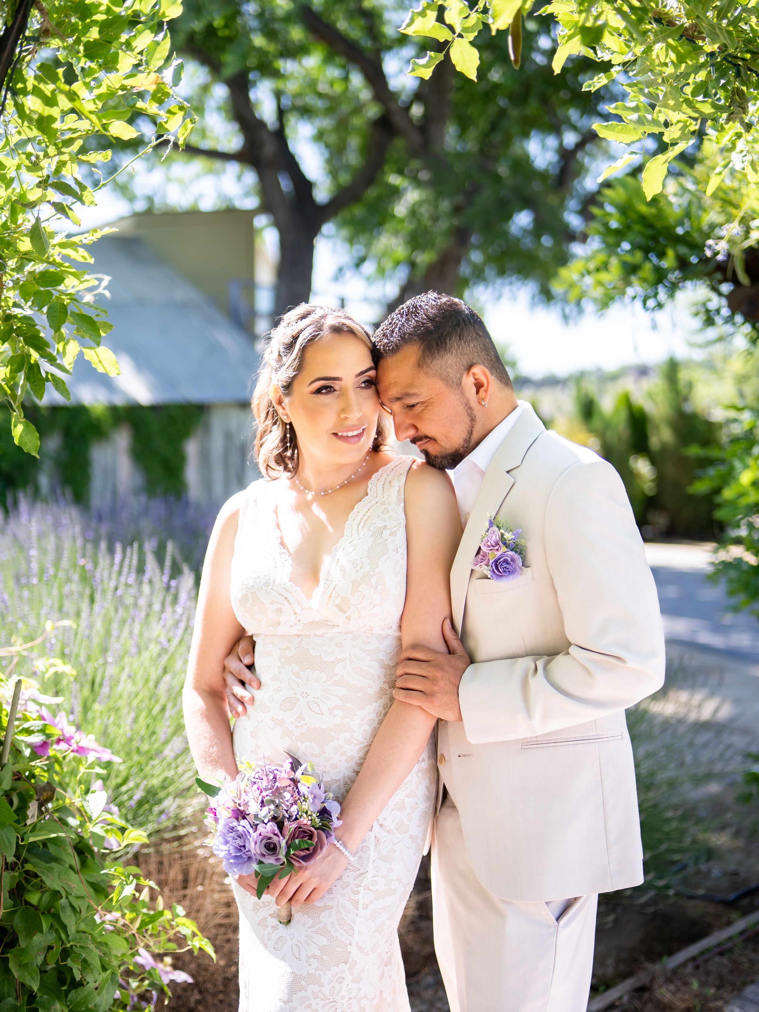 Newlyweds share an intimate moment in a garden with lavender and greenery, wearing cream-colored wedding attire.