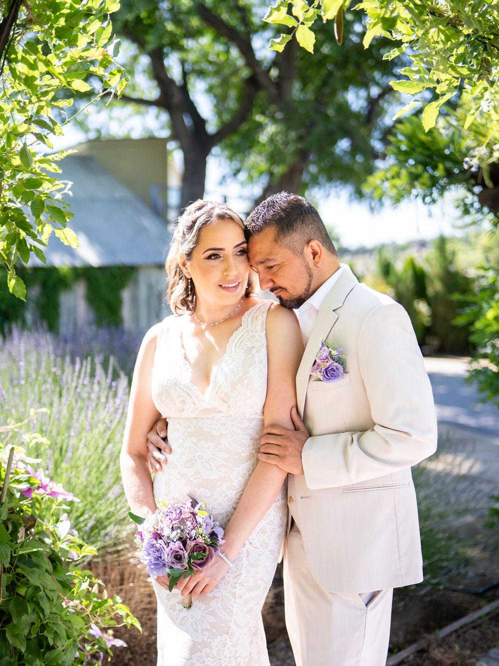 Newlyweds share an intimate moment in a garden with lavender and greenery, wearing cream-colored wedding attire.