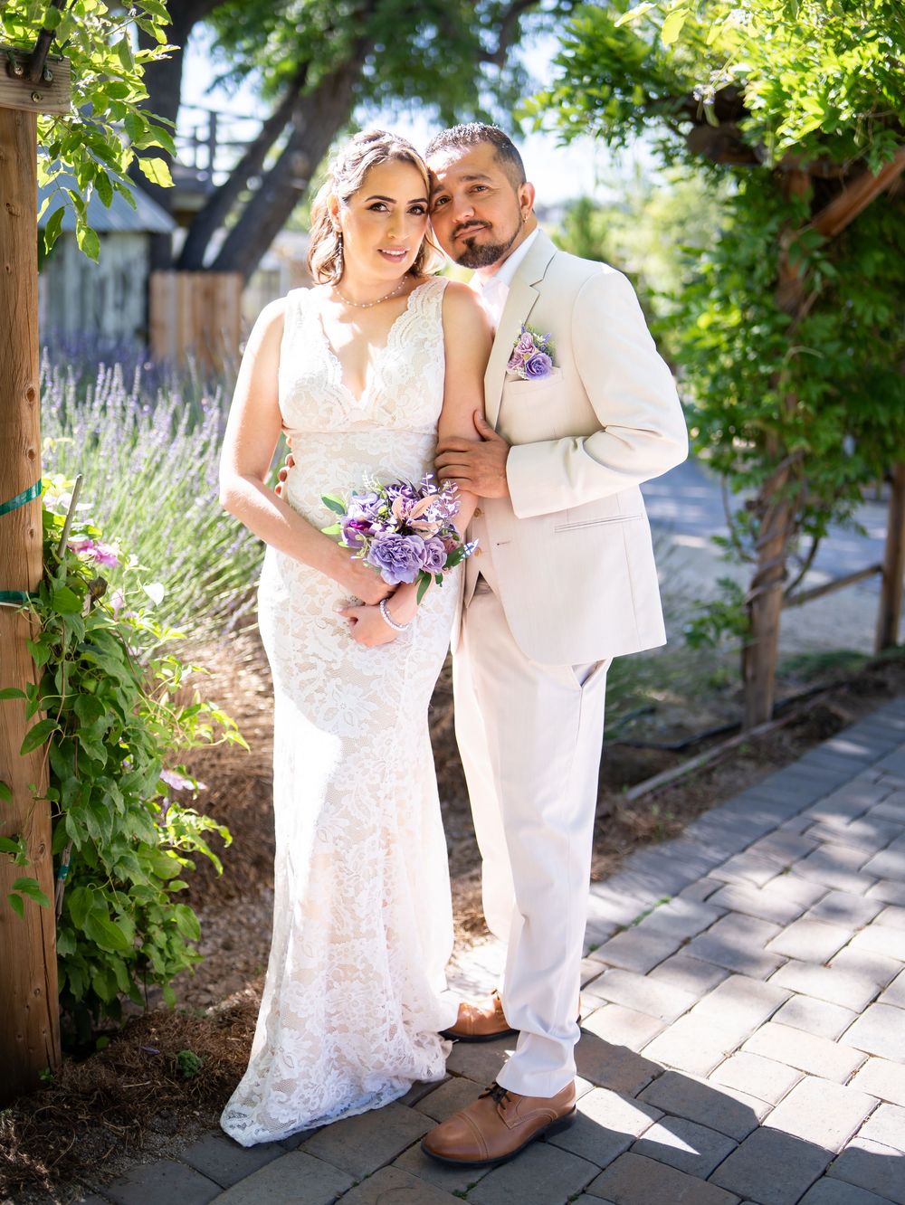 A couple in white wedding attire pose together in a garden setting with purple floral bouquet and lavender plants.