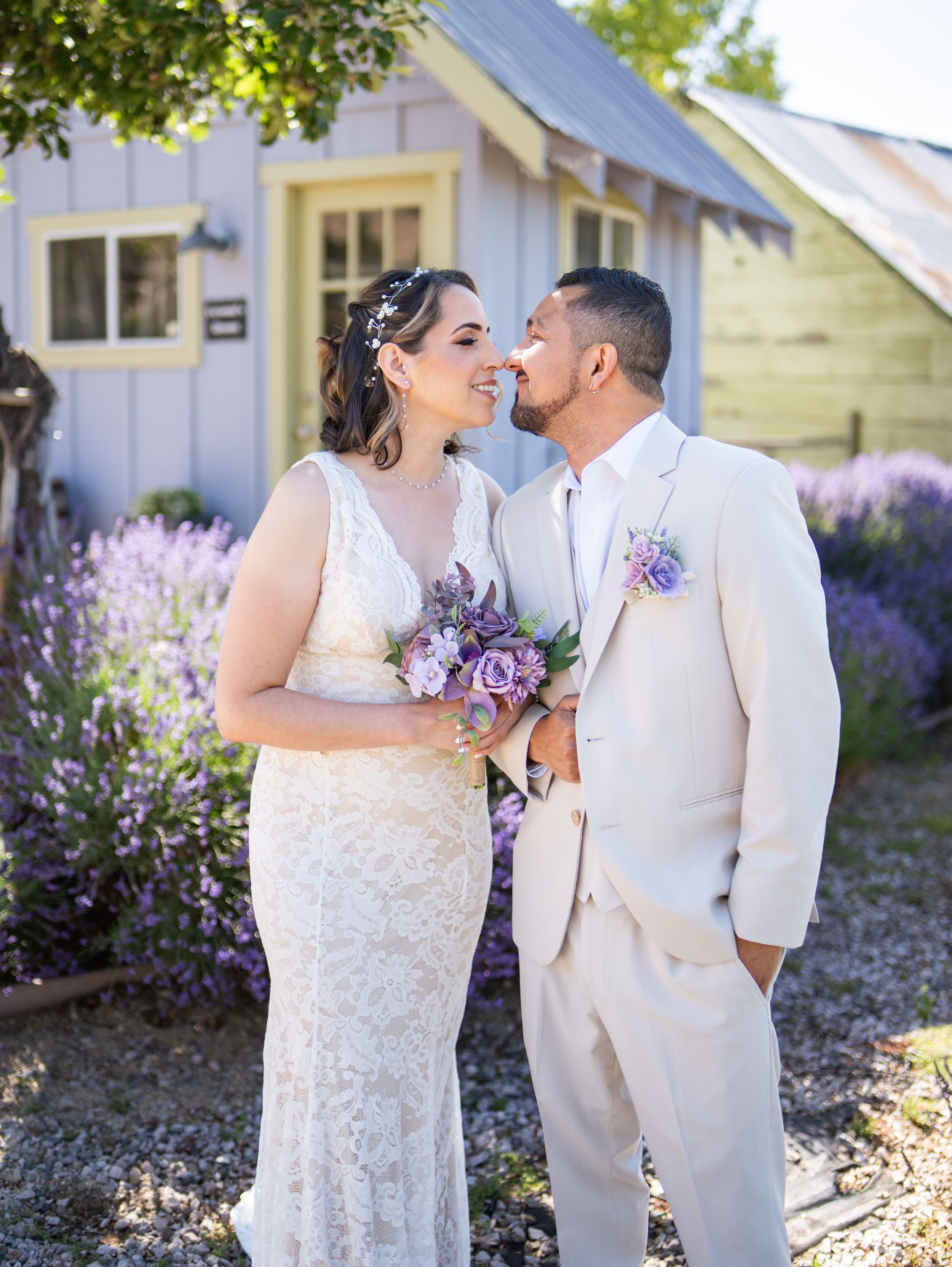 A couple shares a sweet moment in front of a cottage garden with blooming lavender and purple bouquet.