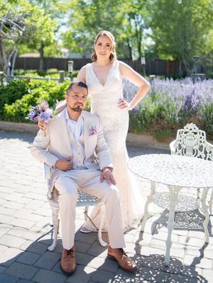 A couple in elegant white attire pose in a garden setting with lavender flowers and ornate patio furniture.