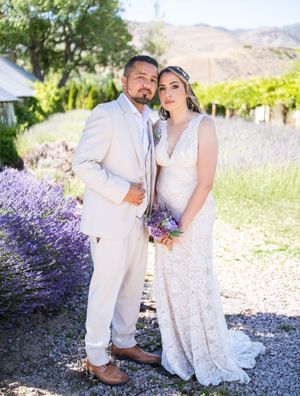A couple poses in a romantic garden setting with blooming lavender and mountains in the background wearing wedding attire.