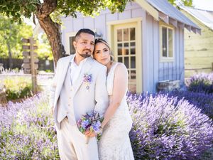 A romantic couple poses in their wedding attire among blooming lavender fields beside a white garden cottage.